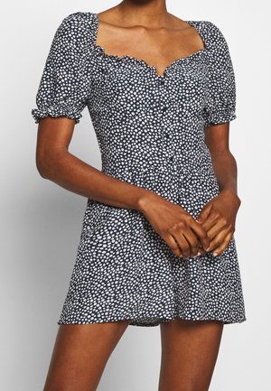 Femme portant une robe courte bleu marine avec imprimé floral blanc, manches bouffantes et encolure carrée à volants, mains jointes devant.