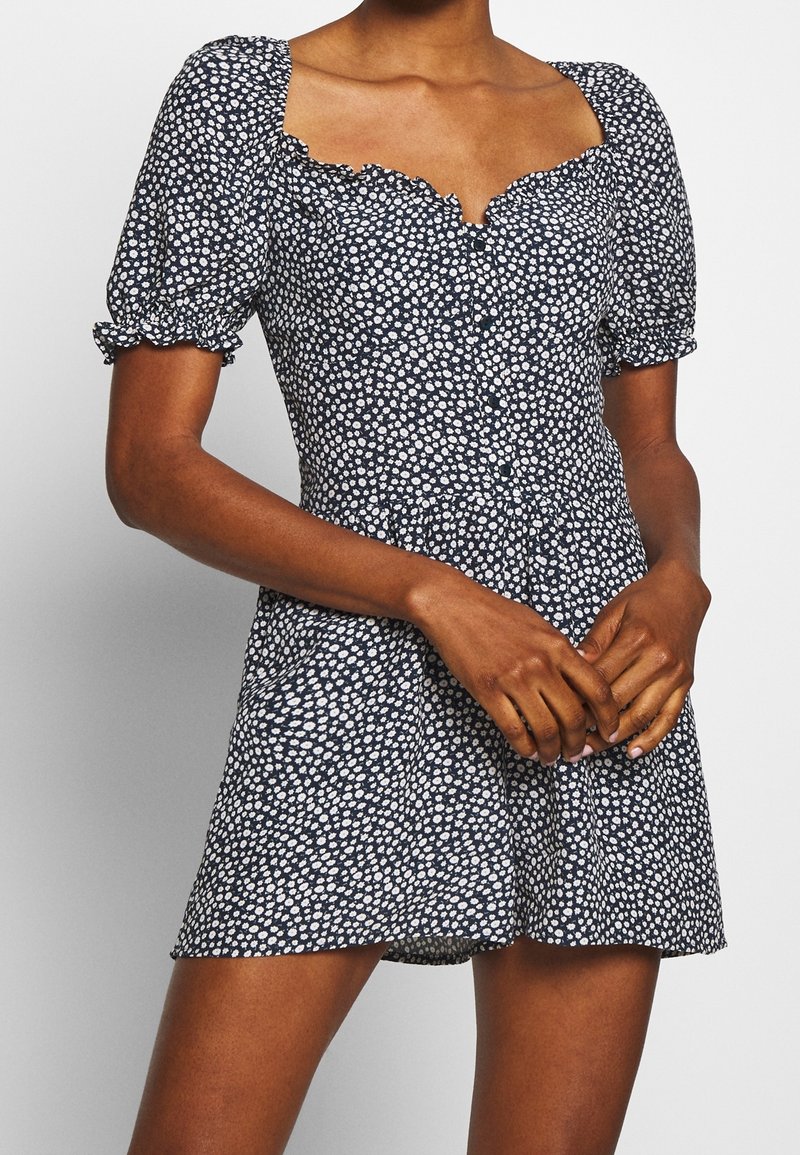 Femme portant une robe courte bleu marine avec imprimé floral blanc, manches bouffantes et encolure carrée à volants, mains jointes devant.