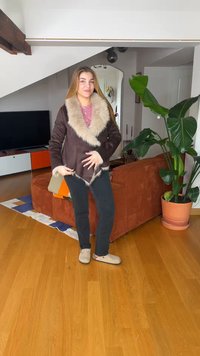 Νεαρή γυναίκα που φορεί καφέ παλτό με γούνα και μαύρο παντελόνι στέκεται σε ξύλινο πάτωμα σε ένα σαλόνι με μεγάλο φυτό και καναπέ.