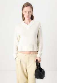 Pull en maille ivoire avec un décolleté en V et un ourlet côtelé, associé à un pantalon beige plissé ; tenant un sac à main en cuir noir.