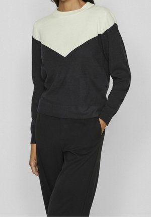 Frau trägt einen langärmeligen schwarz-weißen Pullover mit V-Muster und schwarze Hose, steht mit einer Hand in der Tasche vor einem grauen Hintergrund.