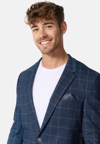 Mørkeblå blazer med ternet mønster, enkelknap lukning og slagkraver. Båret over en hvid t-shirt, med en mønstret lommetørklæde.