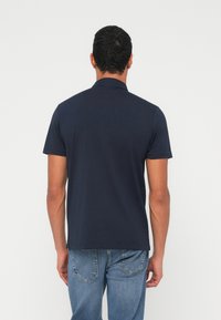 Tumši zils kokvilnas polo krekls ar klasisku apkakli, īsām piedurknēm un sānu atverēm ērtībai, kopā ar gaiši zilām džinsa biksēm.