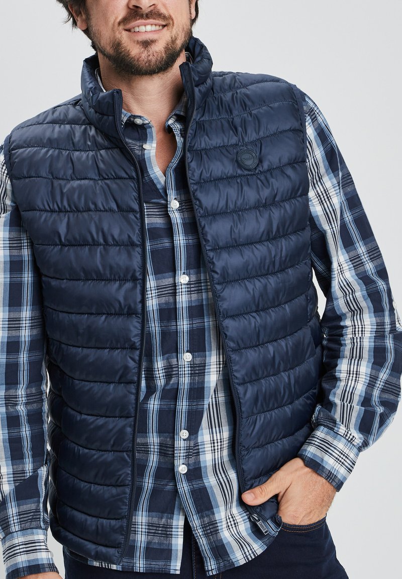 Chaleco de abrigo azul marino con cremallera al frente, que presenta un patrón acolchado. Lleva puesto sobre una camisa de cuadros azul y blanca con botones, mostrando múltiples texturas.