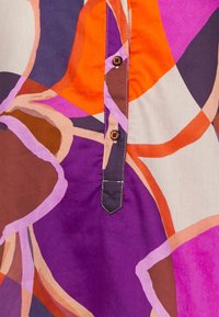 Blusa colorida con formas abstractas en púrpura, naranja y crema. Detalles notables incluyen costuras en contraste y botones marrones.