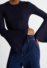 Pull côtelé bleu marine foncé avec des manches évasées porté avec un jean denim bleu foncé taille haute, main glissée dans la poche.