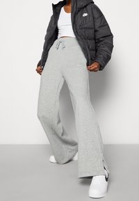 Pantalones de chándal grises, acampanados, con aberturas laterales y cintura con cordón. Combinados con una chaqueta acolchada negra y zapatillas blancas. Logo de Nike visible.