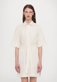 Abito camicia a maniche corte, colore crema chiaro, con righe verticali color beige, chiusura frontale a bottoni e una cintura da annodare in vita. Presenta una tasca sul petto.