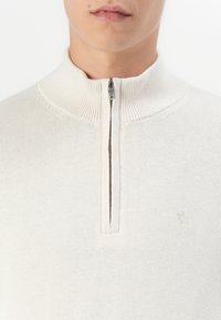 Weißer, Reißverschluss-Pullover mit hohem Kragen, gerippter Textur und kleinem gestickten Logo auf der linken Brust. Weiches Strickmaterial.