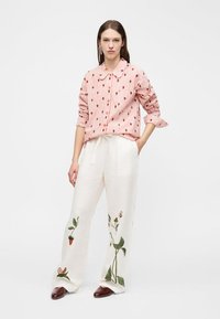 Woman wearing pink and white striped button-up shirt with fruit print, white wide-leg pants with embroidered plants and insects, and brown shoes.