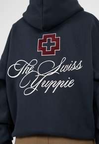 Granatowy sweter z nadrukiem czerwonego krzyża nad napisem "Szwajcarski Yuppie" w białej haftowanej czcionce. Materiał jest miękki i teksturowany.