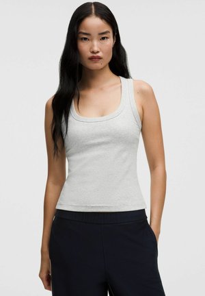 Femme aux longs cheveux noirs porte un haut gris clair sans manches et un pantalon foncé, debout devant un fond clair uni.
