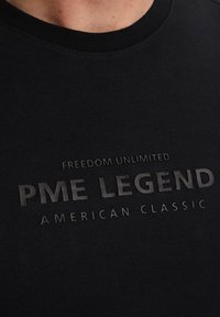 Musta puuvillane T-särk, millel on ümar kaelus ja ülestõstetud tekst: "VABADUS PIIRAMATU PME LEGEND AMERICAN CLASSIC" heledamas varjus.