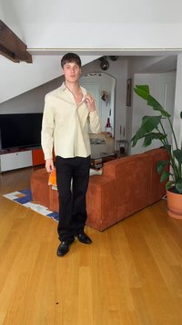 Νέος άνδρας που ρυθμίζει πουκάμισο με μπεζ ρίγες σε σαλόνι με πορτοκαλί καναπέ, μεγάλο γλαστράρι, ξύλινο πάτωμα και τηλεόραση με μοντέρνα έπιπλα.