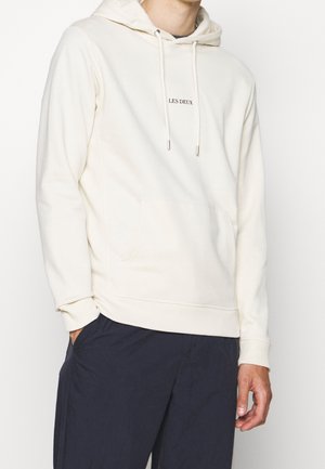 Mand iført en cremefarvet hættetrøje med teksten "LES DEUX" centreret på brystet og mørkeblå bukser, hænder afslappet langs siderne.