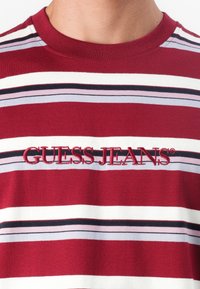 Raudona ir balta dryžuota marškinė su siuvinėtu "GUESS JEANS" logotipu. Minkšta medvilnė, apvalios iškirptės dizainas, su juodais ir levandų dryžiais.
