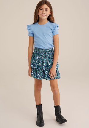 Lichtblauw kortemouwen shirt met ruches, gecombineerd met een meerlagige rok met bloemenprint in blauw, groen en wit. Zwarte enkelboots.
