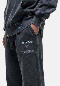 Donkergrijze joggingbroek met zakken, versierd met geschilderde spetterdetails in geel en blauw, en de tekst "GOOD For NOTHING" in contrasterend wit.