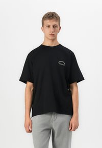 Ung mand iført en sort oversized T-shirt med lille hvid tekst på brystet, parret med lys grå jeans, stående foran en hvid baggrund.