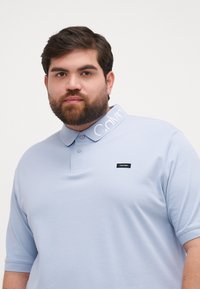 Jasnoniebieska koszulka polo z teksturowanej tkaniny, z kontrastowym białym haftowanym kołnierzykiem i małą czarną metką Calvin Klein na piersi.