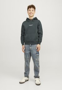 Felpa con cappuccio verde scuro dotata di tasca a marsupio, abbinata a jeans cargo azzurri chiari con più tasche e una vestibilità comoda. Sneakers bianche.