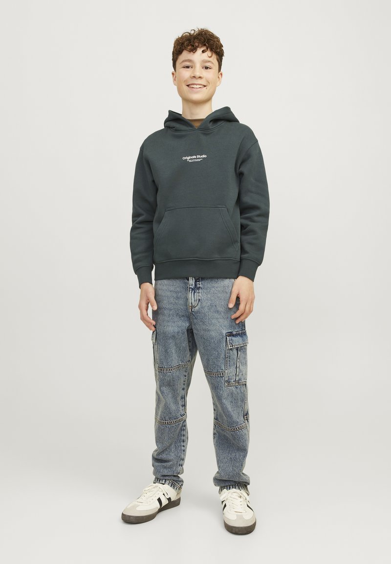 Felpa con cappuccio verde scuro dotata di tasca a marsupio, abbinata a jeans cargo azzurri chiari con più tasche e una vestibilità comoda. Sneakers bianche.