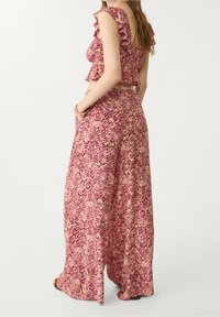 Vrouw in een roze, gestreepte, gerimpelde crop top en bijpassende wijde broek, staande met handen in de zakken, van zich afkijkend.