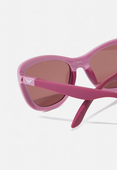 Óculos de sol cor-de-rosa com um acabamento brilhante, armação redonda, lentes castanhas e hastes finas com um pequeno detalhe do logotipo na têmpora.