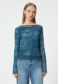 Transparante blauwe top met lange mouwen, overlappende laag en golvende zoom. Kenmerken textpatronen in contrasterende kleuren door het geheel.