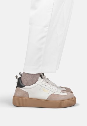 Person trägt beige und weiße Sneakers mit dicken braunen Sohlen, hellbraune Socken und weiße Hosen und steht auf einer weißen Fläche.