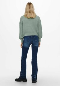 Suéter verde de punto con un corte relajado, mangas abullonadas, combinado con jeans de campana azul oscuro y botas negras de tobillo, visto desde atrás.