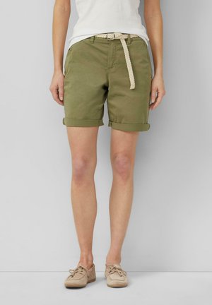Vrouw die opgerolde olijfgroene shorts draagt met een witte gevlochten riem, beige casual schoenen en een wit topje, staand tegen een effen achtergrond.