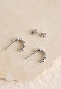 Silberne Creolen mit einem strukturierten Design und erhabenen Akzenten, begleitet von kleineren Ohrsteckern. Die Oberfläche ist glatt und glänzend.