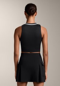 Črn športni top z belim pasom in visokim izrezom, v kombinaciji s črno nabranimi krili, ki imata stranske žepke in gladko teksturo.