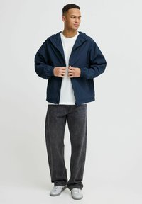 Veste légère à capuche navy avec des poignets élastiques et une fermeture éclair. Associée à un pantalon en denim gris ample et des baskets blanches.