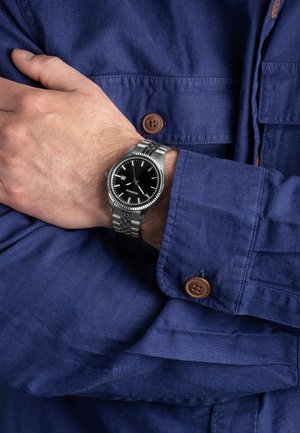 Silberne Uhr mit schwarzem Zifferblatt, Edelstahlarmband und Datumsanzeige, getragen an einem Handgelenk, das mit einem navyblauen Hemd mit braunen Knöpfen gekleidet ist.