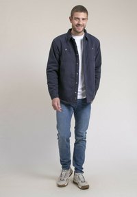 Veste bleu marine à boutons avec poches poitrine, portée sur un t-shirt blanc ; associée à un jean en denim clair et des baskets beiges.