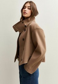 Veste en mélange de laine brun avec un col montant, de grands boutons noirs et des poches latérales. Le design présente une silhouette courte et un style classique.