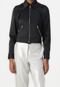Veste courte noire avec col pointu, fermeture éclair et deux poches latérales ; matière lisse avec un design élégant. Portée avec un pantalon argenté brillant.