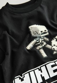 Čierna bavlnená mikina s potlačou pixelových herných postavičiek v bielej a šedej farbe, pod ktorými je výrazný nápis "MINECRAFT". Mäkká textúra.