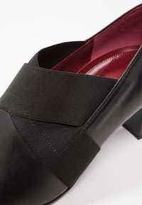Chaussure noire avec une finition en cuir brillant, dotée d'une large bande élastique et d'un intérieur bordeaux. Bout légèrement arrondi et talon épais.