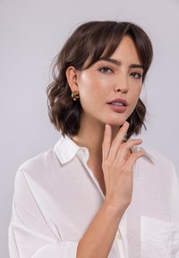 Goldene Creolen, runde Form mit glatter Oberfläche. Das Modell trägt ein weißes Hemd mit Knöpfen, hat ein natürliches Make-up und kurze, wellige Haare.