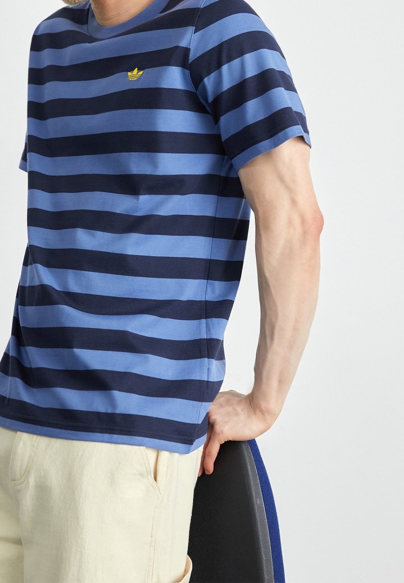 Man i blå och navy horisontellt randig t-shirt med gult logo, beige byxor, luta sig mot en svart och blå stol.