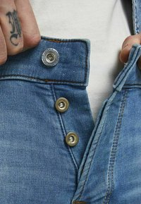 Los jeans de denim azul presentan una cintura con botones metálicos, costuras en contraste y una textura suave. Hay una etiqueta visible en el botón.