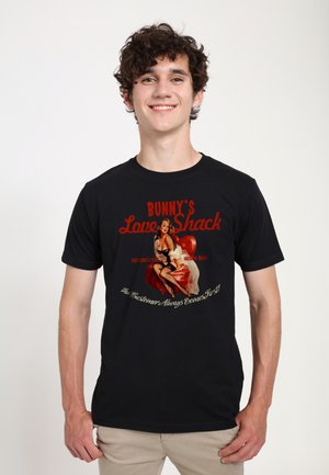 Jonge man die lacht, draagt een zwart T-shirt met een vintage pin-up vrouw zittend op een rode stoel en de tekst "Bunny's Love Shack."