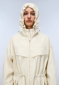 Giacca impermeabile beige chiaro con cappuccio regolabile, vita elastica e tasche frontali. Presenta una chiusura a zip completa e un design minimalista.