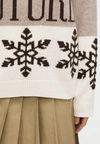 Pull en maille avec un motif beige et blanc cassé, présentant des motifs flocons de neige marron foncé et des poignets côtelés, associé à une jupe plissée kaki.
