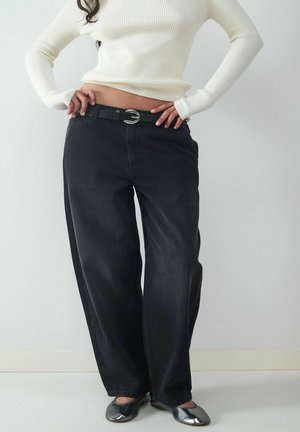 Person in einem weißen gerippten Pullover, hochtaillierten lockeren schwarzen Jeans mit einem schwarzen Gürtel und metallisch silbernen Ballerinas, steht mit Händen in den Hüften.