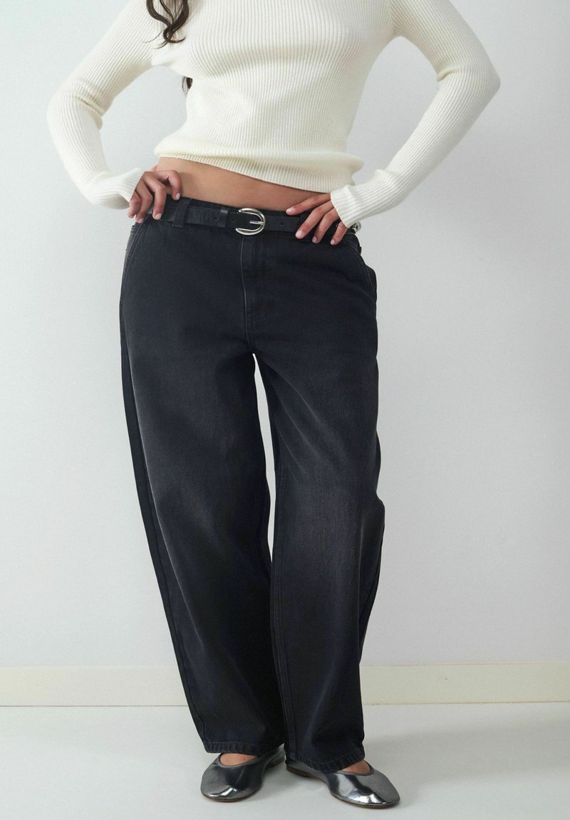 Osoba w białym prążkowanym swetrze, luźnych czarnych dżinsach o wysokim stanem z czarnym paskiem i metalicznych srebrnych balerinach, stoi z rękami na biodrach.