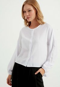 Hvid skjorte med knapper lavet af letvægtsstof, med en rund halsudskæring, lange pufærmer med elastiske manchetter og en cropped kant.